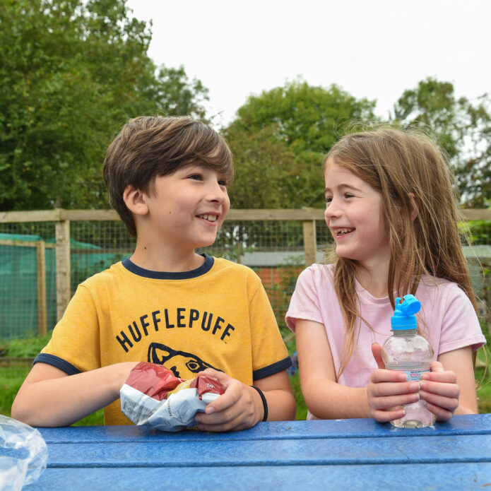 Two children are sitting at a blue picnic table outdoors, smiling and looking at each other. The boy, wearing a yellow shirt with a Hufflepuff logo, holds a sandwich, while the girl, in a light pink shirt, is holding a water bottle. In the background, there are green trees and a fenced area, suggesting a playful outdoor setting.