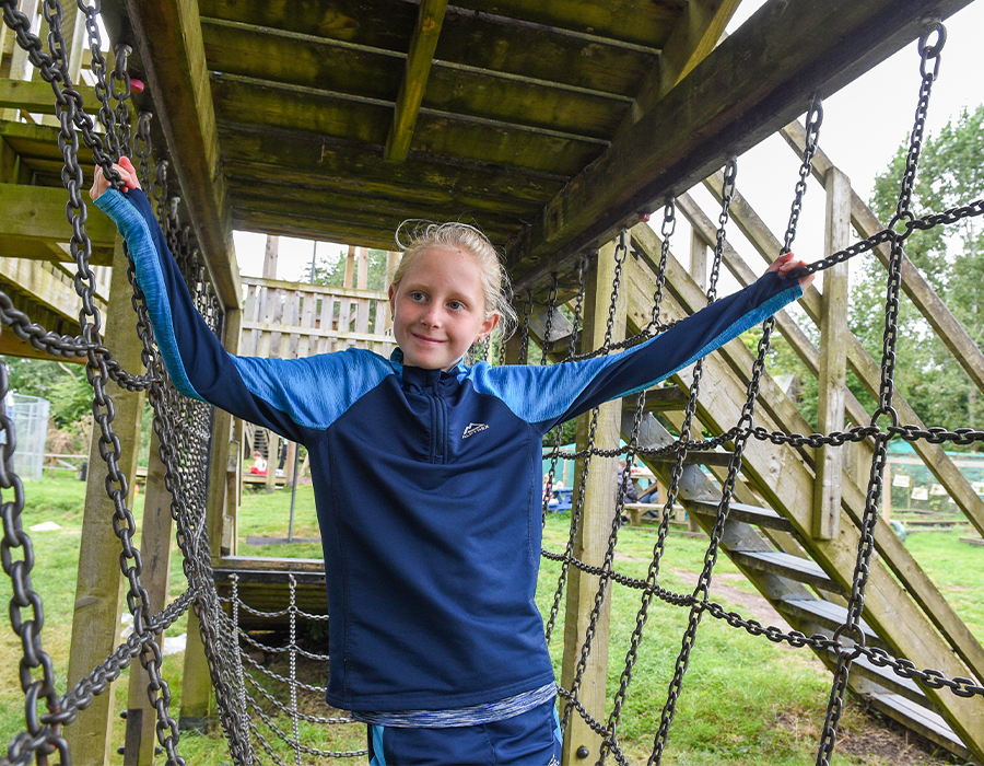 Child in a blue tracksuit balancing on a rope bridge at an outdoor wooden playground surrounded by greenery.
