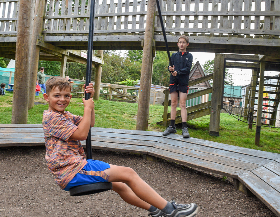 Two children playing on a wooden swing set at an outdoor playground with grassy surroundings.