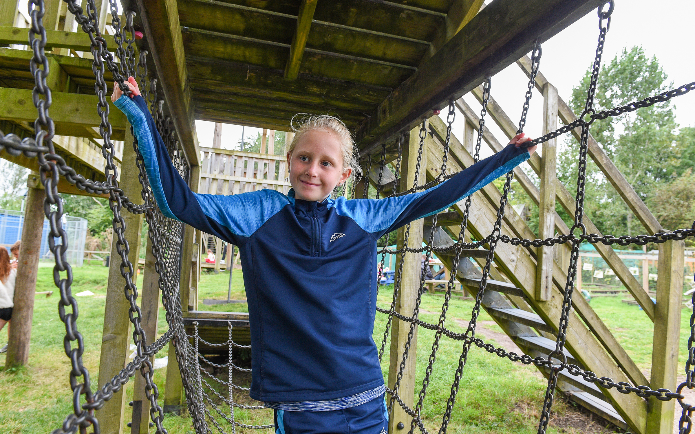 Child in a blue tracksuit balancing on a rope bridge at an outdoor wooden playground surrounded by greenery.