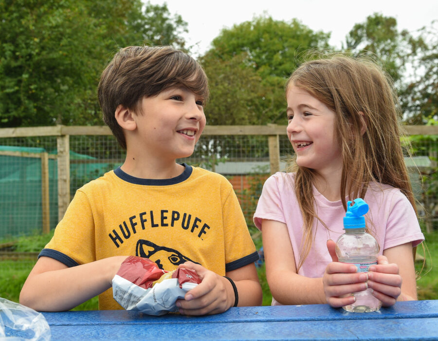 Two children sitting at a blue picnic table outdoors, one in a yellow Hufflepuff shirt with a snack, the other in a pink shirt with a water bottle