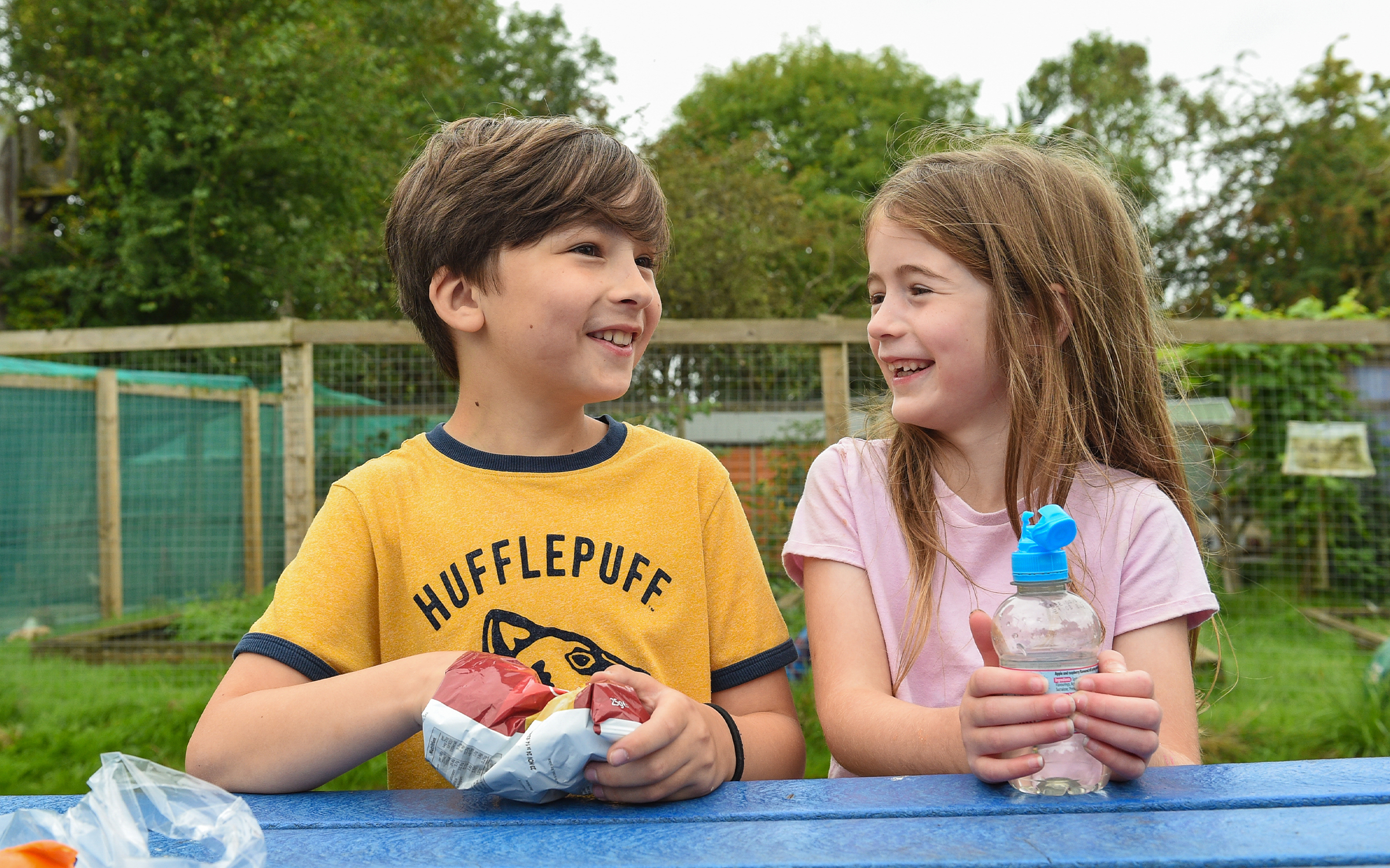 Two children sitting at a blue picnic table outdoors, one in a yellow Hufflepuff shirt with a snack, the other in a pink shirt with a water bottle