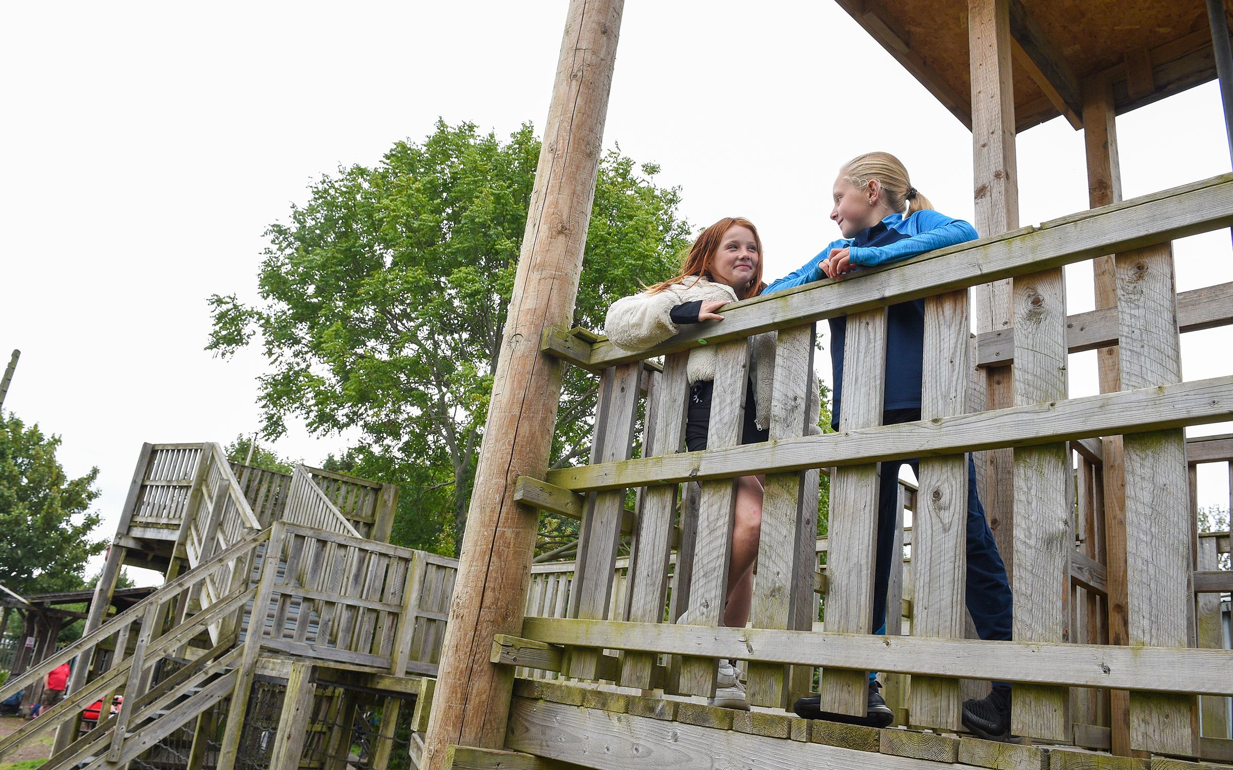 Two children stand on a wooden playground tower, peeking over the railing. A bridge connects the tower to another play area, with trees and more playground equipment in the background.