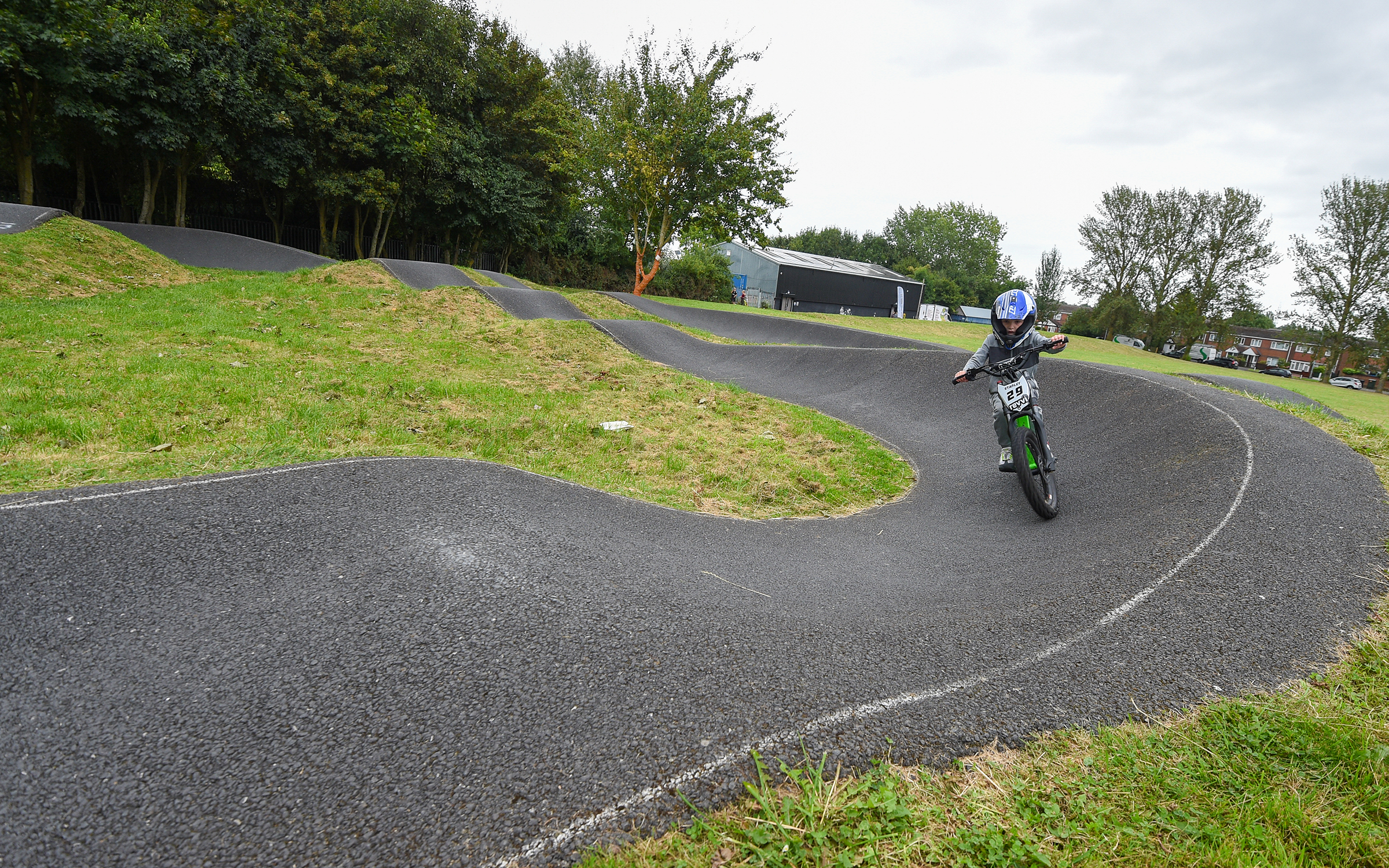 Child riding a small dirt bike on a winding asphalt track surrounded by grass and trees.