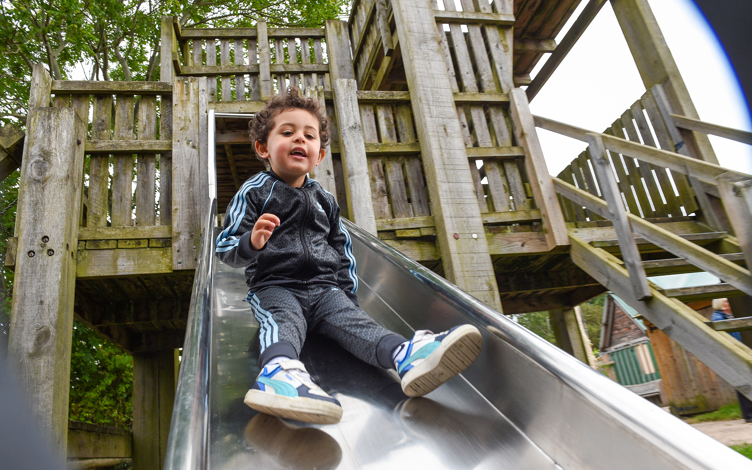 A child in a black tracksuit with blue stripes and blue sneakers slides down a metal slide on a wooden playground surrounded by lush greenery.