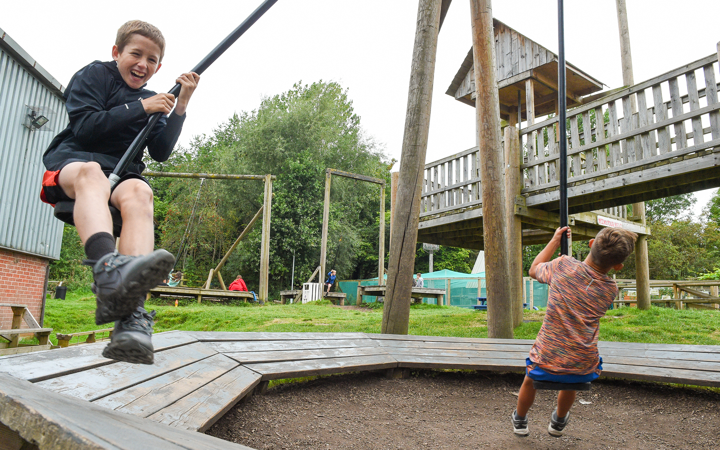Two children playing on a wooden swing set at an outdoor playground with grassy surroundings.