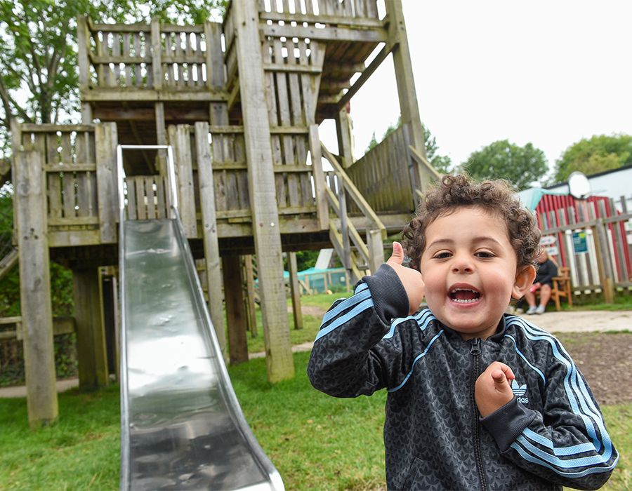 A child in a black tracksuit with blue stripes standing in front of a metal slide on a wooden playground surrounded by lush greenery.