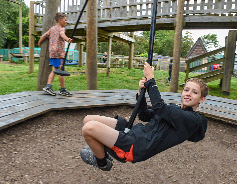 Two children playing on a wooden swing set at an outdoor playground with grassy surroundings.